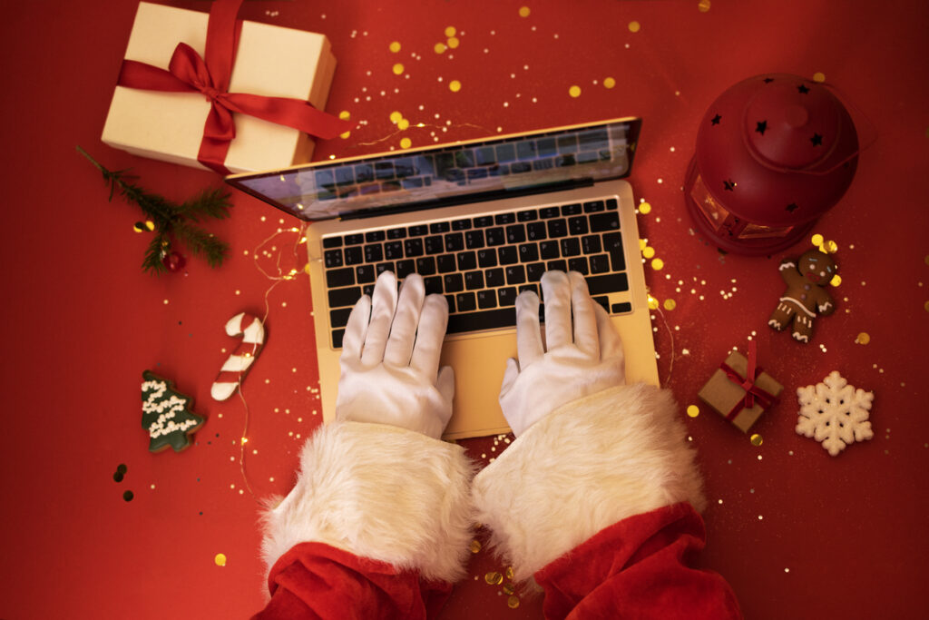 tech gifts Santa Claus with laptop. Hands in white gloves typing on an notebook keyboard. Gift boxes, christmas cookies, lantern and fir branch on red background. Bright backgdrop with sparkles and confetti.