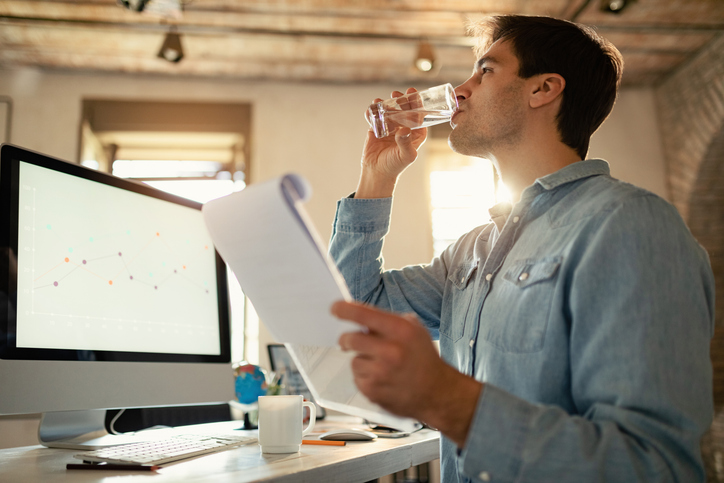 Young freelance worker having a glass of water while working on paperwork in the office. how to stay hydrated at work