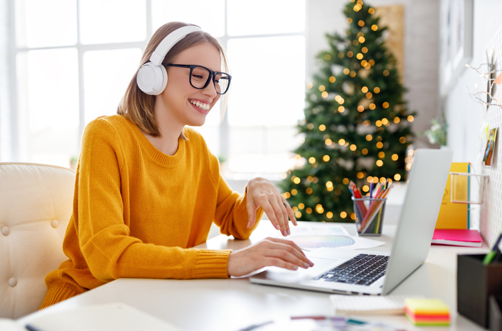 Happy young woman in glasses and headphones smiling and making video call on laptop while sitting at table in home office decorated for Christmas celebration winter office safety checklist